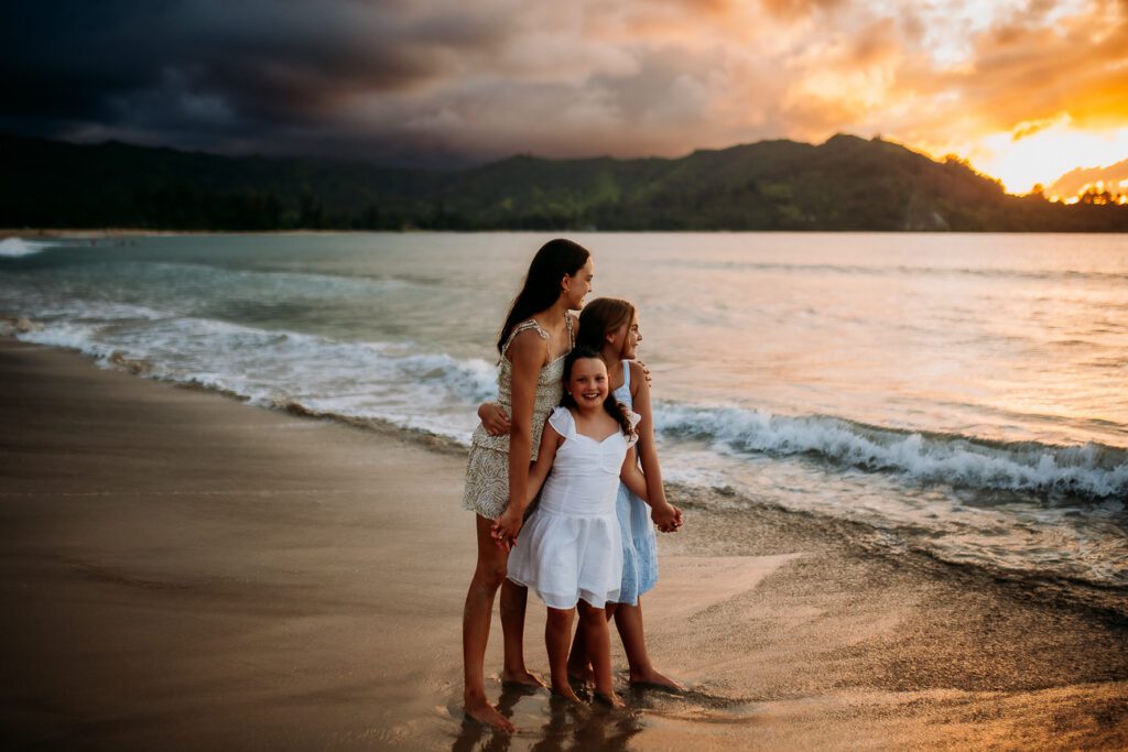 Three girls standing barefoot on a beach at sunset, holding hands and smiling as waves roll in behind them.”, “Three sisters in light dresses on the shoreline at sunset, embracing and looking toward the water.”, “Children posing on the sandy shore during sunset, with dark clouds on the left and warm light on the water.”, “Three girls in white and blue dresses stand together on the beach at sunset, smiling at the camera.”